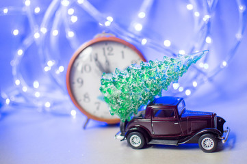 Christmas tree on car toy and clock on bokeh background.Winter.Merry Christmas holiday and Happy New Year concept.The car is in a hurry to deliver the Christmas tree home. Copy space, filter effect.