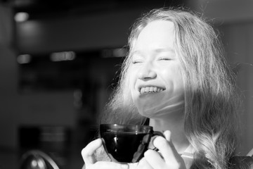 Portrait of a young pretty girl with a cup of coffee sitting in front of the window on a sunny day. Bright sun rays fall on the face. Blurred background. Black and white.