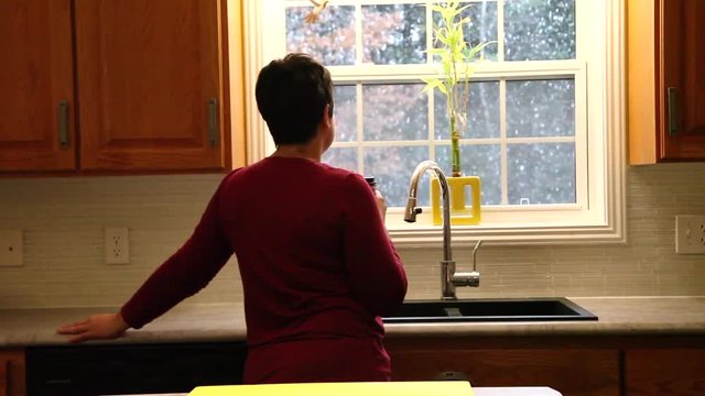  A woman holds a cup of coffee while looking outside at the falling snow from inside her warm home