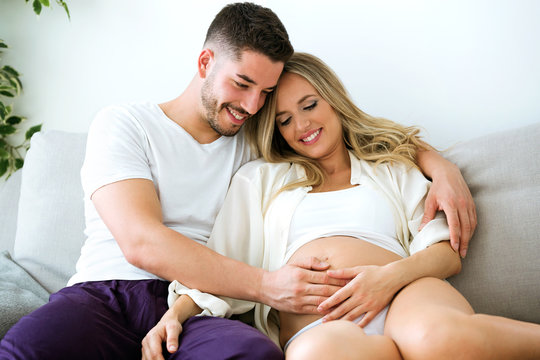 Handsome young man touching belly of beautiful pregnant woman while lying on the sofa at home.