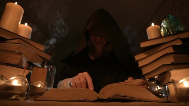 Medium close-up girl magician in a hood in a dark room by candlelight and looking for a spell turning over a book. Low key. Mystic Small DOF