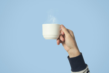 Male hand holding a white cup with hot coffee or tea on a light blue background. Breakfast concept with hot coffee or tea