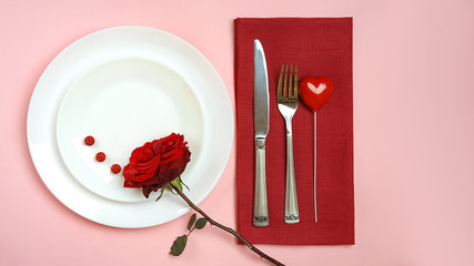 Valentines day table setting with red rose bud, fork and knife heart red tablecloth on pink pastel background. Romantic table setting for Valentines day dinner date or celebration wedding.