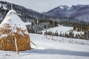 Winter rural landscape, a haystack on the background of snow-capped mountains and forest