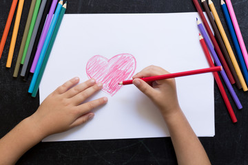 Kid's hands with pencil draws heart on white paper. Mother's day concept. Celebration. Hand made card. Top view.