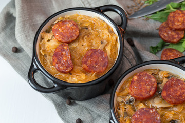 Slovak Christmas national cabbage soup in two small black pots with sausage on the tablecloth background.