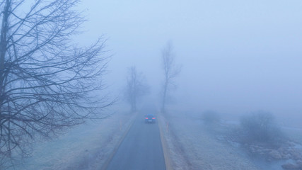 Obraz premium AERIAL: Car driving along scary foggy country road leading through misty fields