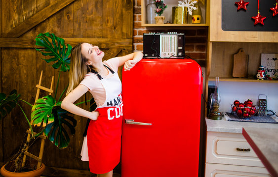 The sweet girl in an apron is in the kitchen near the red refrigerator