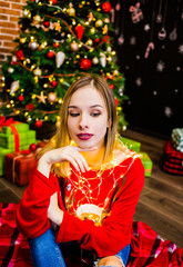 A exquisite girl in a red sweater sitting at a tree and holding garlands in her hands.