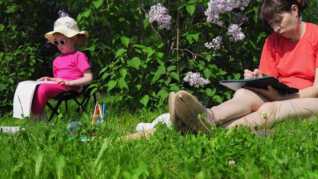 A young mother with dark hair and a cute little daughter paint next to a blooming lilac in the park on a spring sunny day, camera movement from right to left.