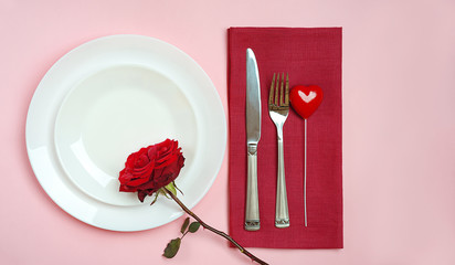 Valentines day table setting with red rose bud, fork and knife heart red tablecloth on pink pastel background. Romantic table setting for Valentines day dinner date or celebration wedding.