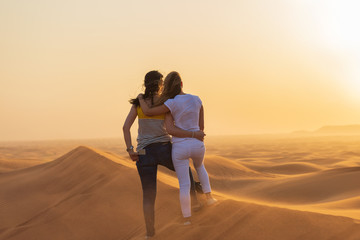 Fototapeta na wymiar Couple of young tourist women looking at sunset in sand dunes in Dubai desert. Beautiful view for two friends of desert and sunset from top of sand dunes