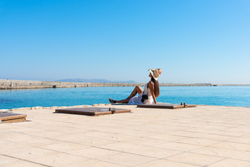 Happy tourist girl on holiday trip to Heraklion, Crete, Greece. Young woman traveler enjoyes sunshine sitting on the dock of Heraklion Venetian port marina. Dragon Island at background.