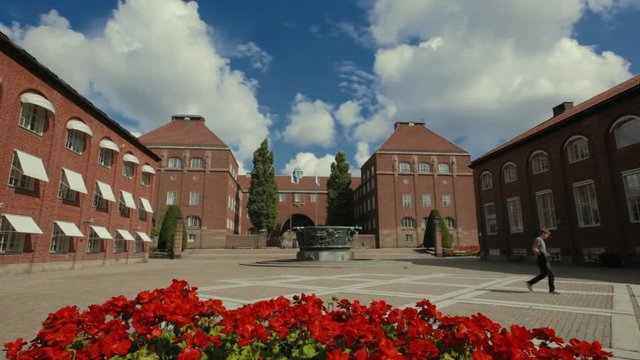 A student walks across a wide courtyard between buildings just inside the entrance to KTH University. A large water feature in the center of the space. Bright red flowers in the foreground.