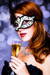 Style redhead girl in dark dress and mask with glass of drink on gray background with bokeh