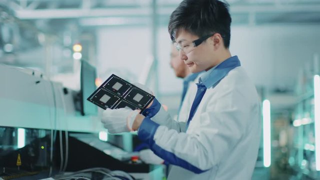 High-Tech Factory: Quality Control Engineer Checks Electronic Printed Circuit Board it for Damages. In the Background Assembly Line for PCB with Surface Mount Pick and Place Technology.