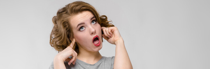 Charming young girl in a gray T-shirt on a gray background. The girl covered her ears with her...