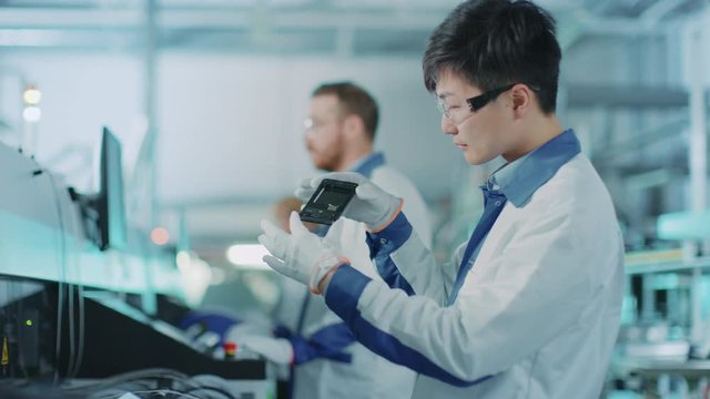 High-Tech Factory: Quality Control Engineer Picks up and Visually Checks Smart Electronic Device for Possible Damages. In Background Assembly Line for PCB with Surface Mount Pick and Place Technology.