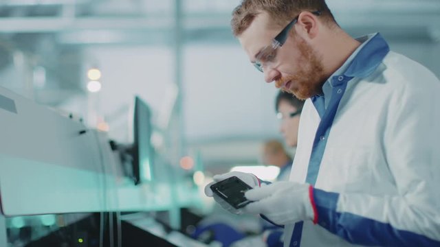 High-Tech Factory: Quality Control Engineer Picks up, Visually Checks Smart Electronic Device for Possible Damages. In the Background Assembly Line for PCB with Surface Mount Pick and Place Technology