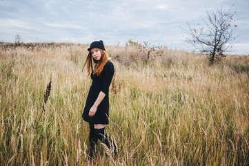 Naklejka na ściany i meble beautiful red-haired woman in a black dress walks on an autumn field.