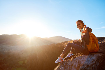 first view of woman hiker with backpack sits on edge of cliff against background of sunrise and extends a hand - obrazy, fototapety, plakaty