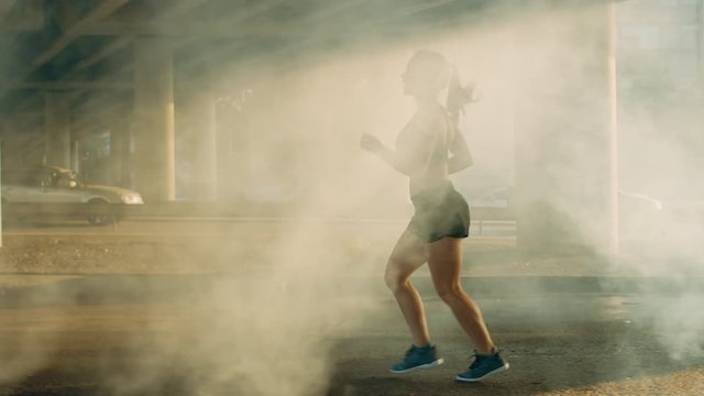 Slow Motion Shot of a Beautiful Fitness Girl in Black Athletic Top and Shorts Jogging Through a Smoky Street. She is Running in an Urban Environment Under a Bridge with Cars in the Background.