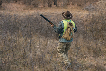 A hunter in a hat with a gun in camouflage and a reflective vest in the steppe