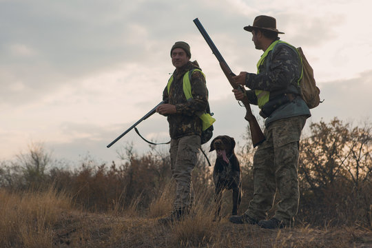 Two Hunters With Guns And A Dog On A Hill Against The Blue Sky