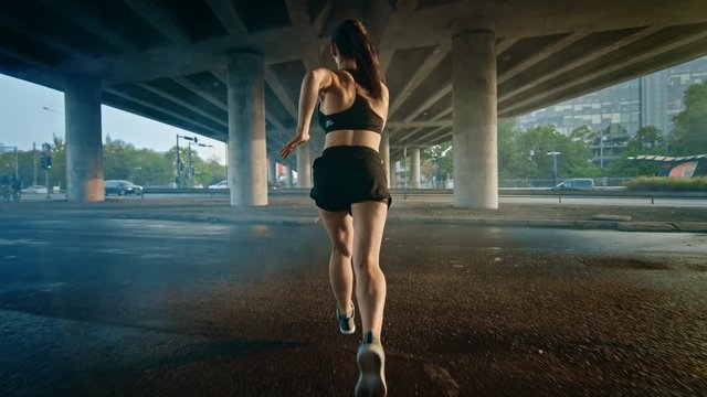 Backshot of a Strong Fitness Girl in Black Athletic Top and Shorts Jogging Through a Smoky Street. She is Running in an Urban Environment Under a Bridge with Cars in the Background.