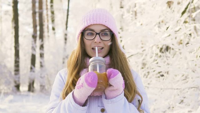 Beautiful young blonde woman in glasses pink hat, scarf, mittens, white jacket enjoying hot mulled wine beverage outside on winter snowy day.
