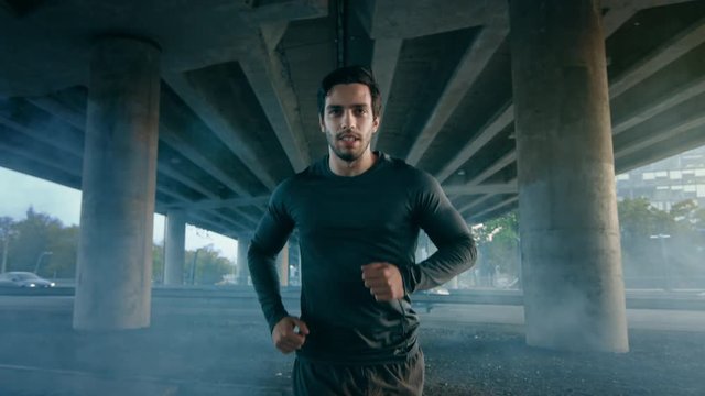 Close Up Shot of a Tired Athletic Muscular Young Man in Sports Outfit Jogging in the Street. He is Running in an Urban Environment Under a Bridge with Cars in the Background.