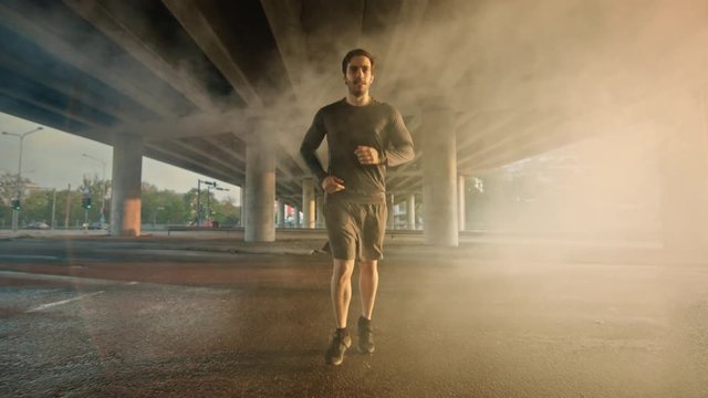Athletic Muscular Young Man in Sports Outfit Jogging in a Street Filled With Steam. He is Running in an Urban Environment Under a Brindge with Cars in the Background.