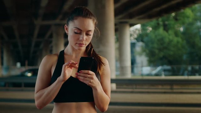 Beautiful Strong Fitness Girl in Black Athletic Top is Using a Smartphone and Smiling on a Street. She is in an Urban Environment Under a Bridge with Cars in the Background.