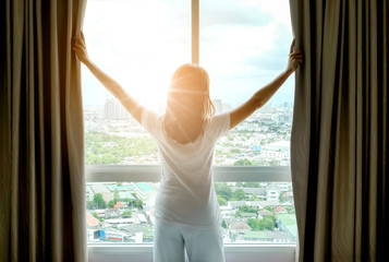 Morning of a new day woman wake up in the bedroom with her refreshing. She stand at the window , raised her two arms and look city view get sunlight - obrazy, fototapety, plakaty