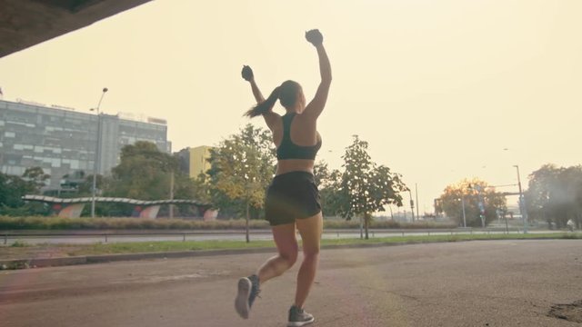 Backshot of a Strong Fitness Girl in Black Athletic Top and Shorts Jogging on a Street. She is Celebrating the End of Her Workout. Athlete is Running in an Urban Environment Under a Bridge with Cars.