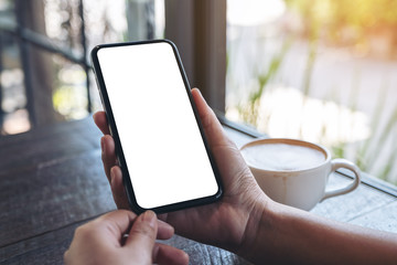 Mockup image of hands holding black mobile phone with blank white desktop screen with coffee cup on wooden table in cafe