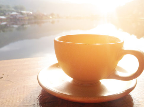 Selective focus picture of a cup of coffee on wooden table with river in the morning sunrise