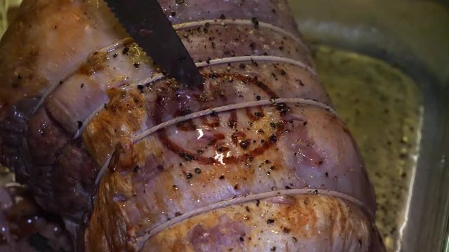 Slow motion of stabbing a half done pork joint with a serrated kitchen knife in a glass cooking dish.