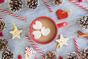 Hot cocoa in red mug with heart marshmallows surrounded by handmade ornaments, candy canes, and pinecones