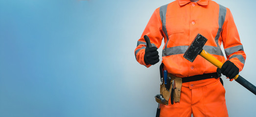 Builder worker holding in hand a big sledgehammer and is showing a thumbs up isolated on blue...