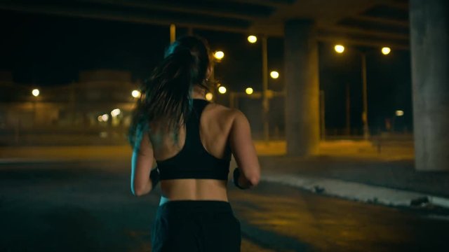 Beautiful Fitness Girl in Black Athletic Top and Shorts is Jogging on the Street. She is Doing a Workout in a Night Urban Environment Under a Bridge with Cars in the Background.