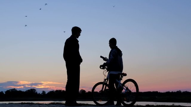 The guy picks up the phone and takes a selfie on the phone at dawn. Silhouette of a man and a teenager with a bicycle being photographed against the background of the morning sky. A happy family.