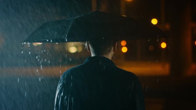 Backshot of a Young Caucasian Shorthaired Man, Wearing a Jeans Coat Walking in the Rain Under an Umbrella. He's Walking at Night in the City.