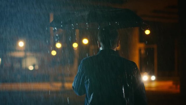 Backshot of a Young Caucasian Shorthaired Man, Wearing a Jeans Coat Walking in the Rain Under an Umbrella. He's Walking at Night in the City.