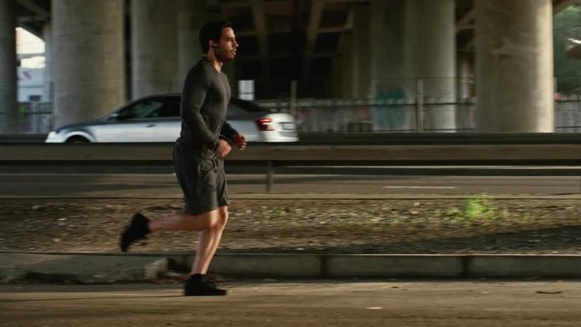 Athletic Young Man in Earphones and Sports Outfit is Jogging in the Street. He is Running in an Urban Environment Under a Brindge with Cars in the Background.