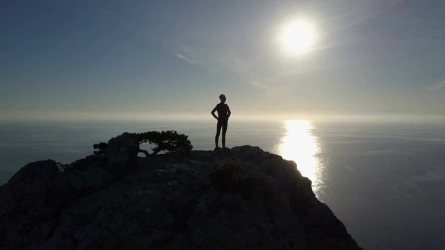Aerial silhouette of young woman standing on the top of a mountain facing the sea. Lady on the summit in beautiful scenery.