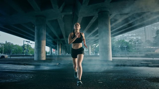 Portrait Shot of a Beautiful Confident Fitness Girl in Black Athletic Top and Shorts Jogging Through a Smoky Street. She is Running in an Urban Environment Under a Bridge with Cars in the Background.
