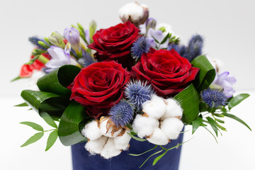 A bright bouquet of red flowers (rose and anthurium) with green leaves, Alstroemeria and cotton flowers in a blue box for hats on a light background