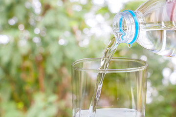Pouring of clear drink water from bottle into the glass on blurred green nature background with copy space for text.