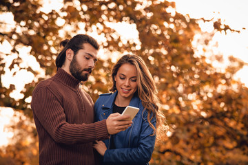 Young couple using cellphone in autumn colored park.